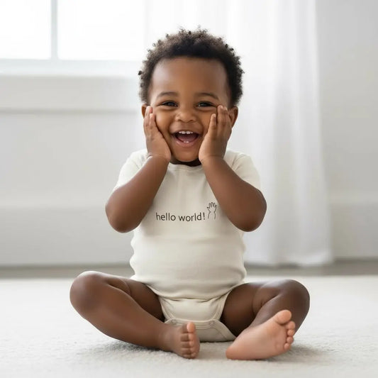 Toddler wearing a natural cream 'hello world!' onesie sitting on a light-colored floor