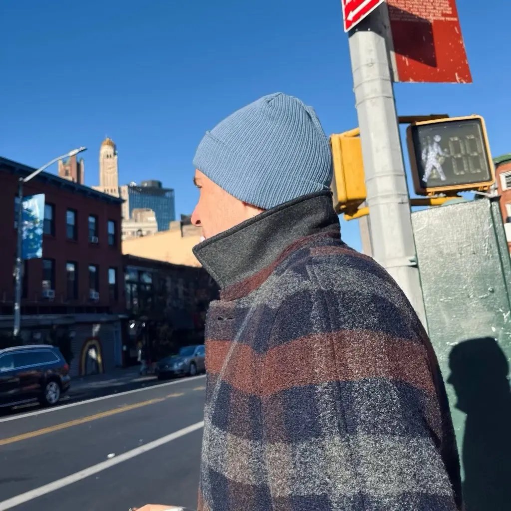 Person wearing a blue beanie and plaid coat standing on a city street with traffic lights and buildings in the background.