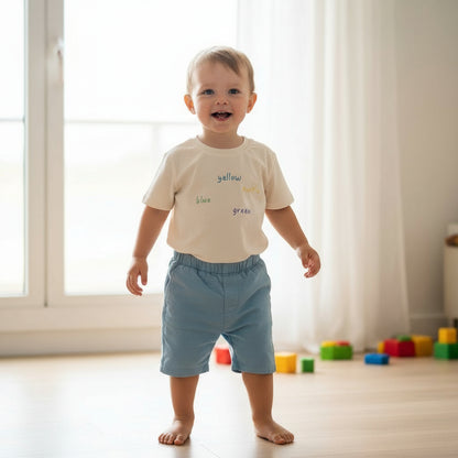 Toddler wearing an oat cream t-shirt with text and light blue shorts standing in a room with colorful blocks on the floor