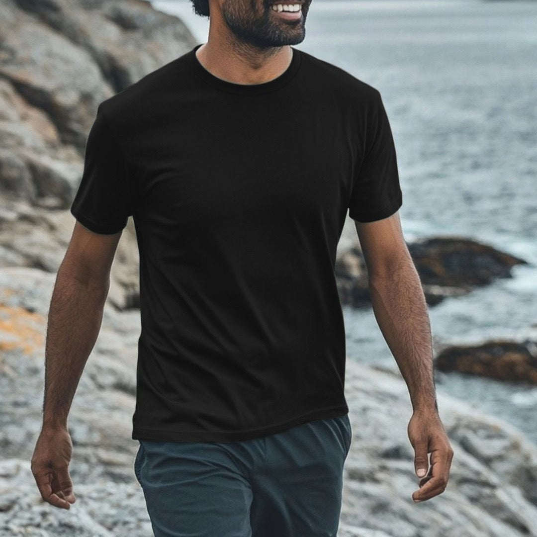Man wearing a black t-shirt and gray shorts walking along a rocky shoreline.