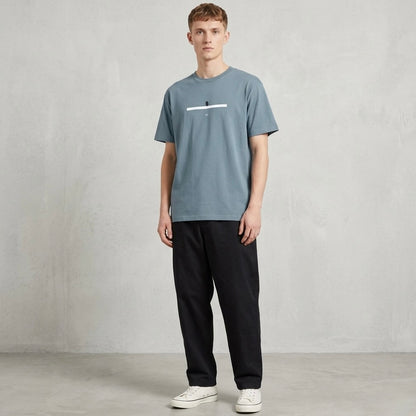 Man wearing a stone blue t-shirt and black pants against a plain background