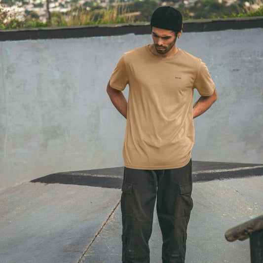 Man wearing a beige t-shirt and dark pants standing in an outdoor setting.