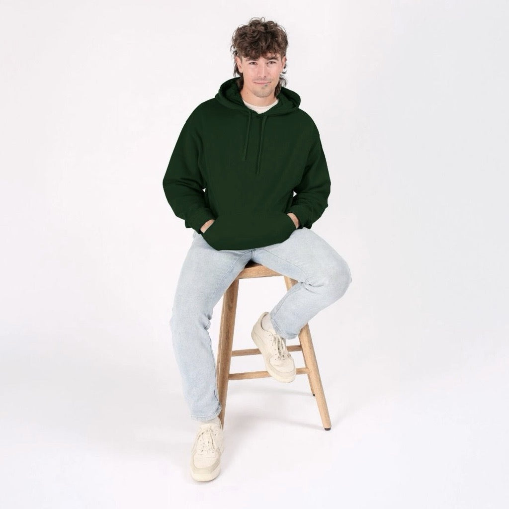 Person wearing a green hoodie and light gray pants sitting on a wooden stool against a white background