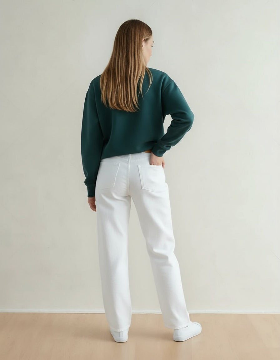 Person wearing a green sweatshirt and white pants standing against a plain wall.